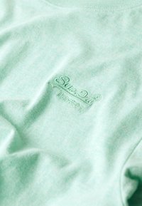 T-shirt in cotone verde menta con una texture morbida, caratterizzato da un logo ricamato di un verde più chiaro sul davanti. Collo girocollo casual.