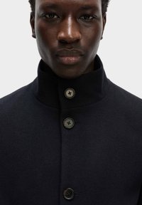 Marineblå uldfrakke med høj krave, der har tre sorte knapper og en glat tekstur, designet til en moderne pasform.