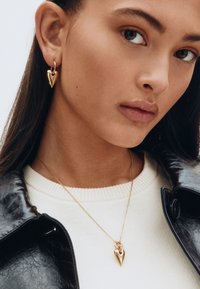 Guldörhängen i hjärtform och matchande halsband med kedja, placerade mot en vit stickad topp och svart läderjacka.