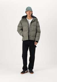 Olive green puffer jacket with a hood, front snap closure, gray lining, and two chest pockets. Paired with black pants and black shoes.