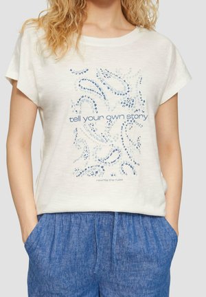Vrouw draagt een wit shirt met korte mouwen met een abstract blauw patroon en tekst "tell your own story" en een blauwe broek met zakken.