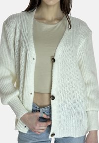 Witte gebreide cardigan met een gestructureerd patroon, voorzien van een V-hals en drie donkere knopen. Draagt over een beige top en lichte spijkerbroek.