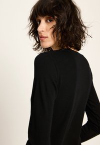 Frau mit welligem dunklem Haar, die ein schwarzes langärmliges Oberteil trägt und über ihre Schulter gegen einen schlichten hellen Hintergrund schaut.