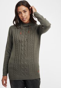 Pull en maille torsadée vert olive avec un col montant, des cordons de serrage réglables et un patch en cuir au bas. Poignets et ourlet côtelés.