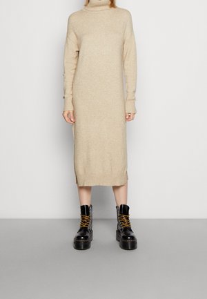 Abito a collo alto beige con maniche lunghe, polsini a coste, spacchi laterali, abbinato a stivali platform neri con lacci gialli.