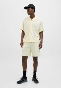 Camisa de manga corta y pantalones cortos amarillos claros a juego con un patrón de olas. El conjunto tiene un corte relajado y se combina con zapatillas negras y calcetines.