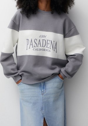 Personne portant un sweat-shirt gris et blanc avec le texte "1991 Pasadena California" et une jupe en jean bleu clair avec une fente à l'avant.