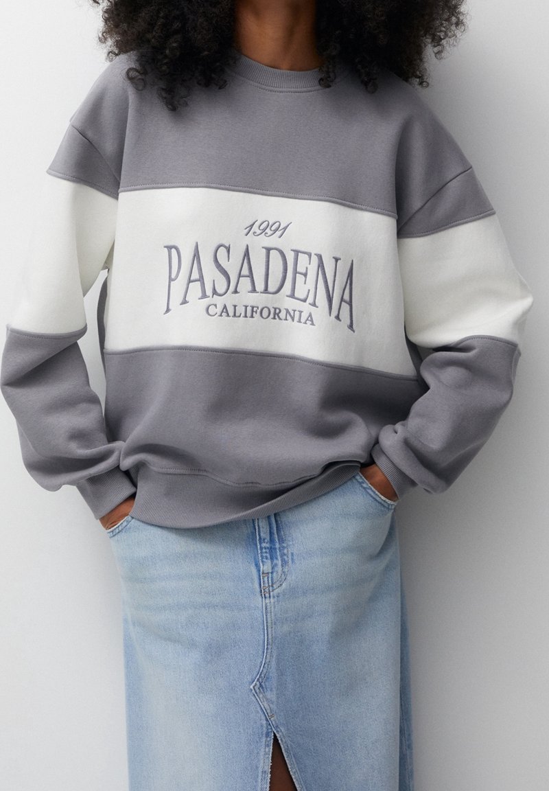 Personne portant un sweat-shirt gris et blanc avec le texte "1991 Pasadena California" et une jupe en jean bleu clair avec une fente à l'avant.