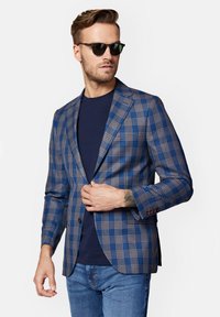 Niebieska marynarka w kratę z brązowymi akcentami, dopasowany krój, klapy notch, zapięcie na jeden guzik, noszona na granatowej koszuli, w zestawie z dżinsami.