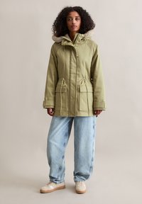Groene parka met een fleece-voering in de capuchon, voorzien van een trekkoord in de taille, twee voorkruiken en knoopsluitingen. Gecombineerd met lichtblauwe baggy jeans en sneakers.