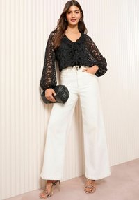 Blouse en dentelle noire avec des manches larges, associée à un pantalon taille haute à jambes larges blanc. Tient une pochette noire texturée, portant des talons nus.