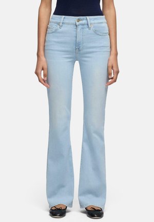 Kobieta w jasnoniebieskich dzwonowatych jeansach z wysokim stanem, z przednimi kieszeniami i czarnych balerinach stoi na białym tle.