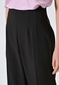Pantaloni neri a vita alta con pieghe e una texture liscia. Presentano una tasca laterale e una vita aderente. Il tessuto sembra leggero e fluido.