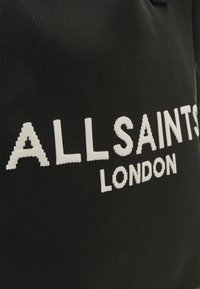 Tissu noir avec texte brodé blanc "ALLSAINTS LONDON" en lettres majuscules grasses.
