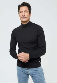 Pull à col roulé noir en tissu tricoté, coupe ajustée, texture lisse, avec un logo discret sur l'ourlet, associé à un jean bleu clair.