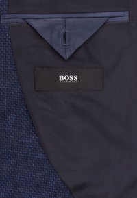 Marinblå jacka med inredning i texturerat tyg och slät foder. Har ett triangulärt ficklock och en etikett med "BOSS Hugo Boss."