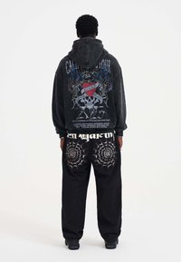 Black hooded sweatshirt with graphic print featuring skulls and roses on back; paired with black pants adorned with spiderweb embroidery.