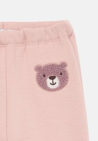 Pink bomuldsbukser med elastisk talje, prydet med et broderet ansigt af en fluffy bjørn på forsiden, der fremhæver teksturer og subtile detaljer.