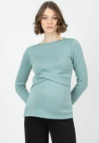 Långärmad top i teal-färg gjort av mjukt tyg, med en vriden framsida och båthalsringning, tillsammans med svarta byxor.