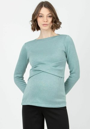 Langärmliges Top in Teal, aus weichem Stoff gefertigt, mit einem gedrehten Vorderteil und einem Bootsausschnitt, kombiniert mit schwarzen Hosen.