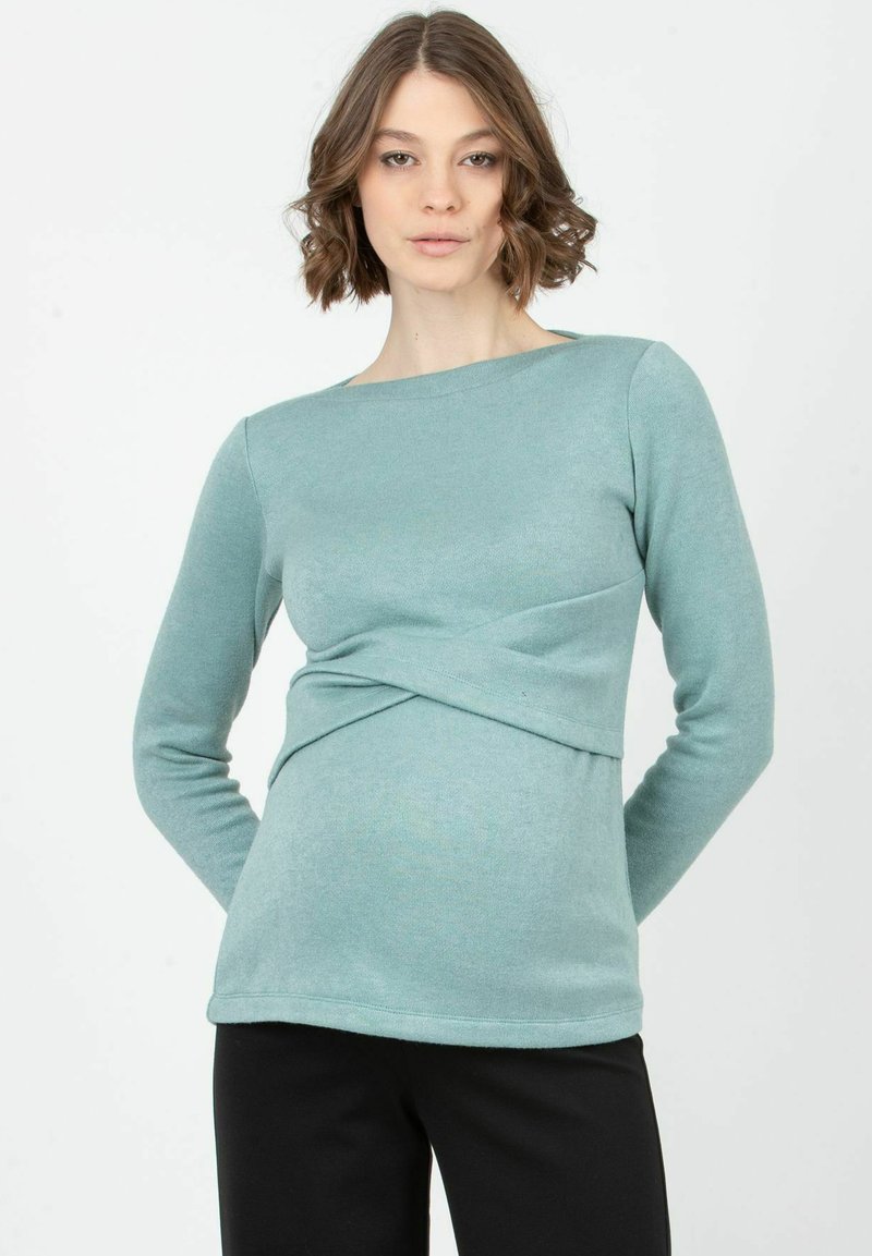 Långärmad top i teal-färg gjort av mjukt tyg, med en vriden framsida och båthalsringning, tillsammans med svarta byxor.