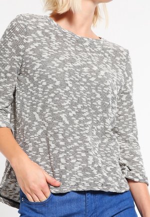 Kvinde iført en sort-hvid, horisontalt stribet, langærmet trøje med rund halsudskæring og blå jeans, hånd i lommen.