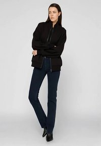 Černá bomber bunda se zipem vpředu, žebrovanými manžetami a lemem; v kombinaci s tmavě modrými úzkými džíny a černými špičatými kotníkovými botami.