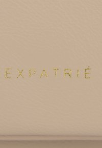 Surface en cuir beige avec le mot « EXPATRIÉ » embossé en petites lettres dorées au centre.