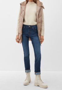 Beige vadderad väst, krämfärgad texturerad tröja, mörkblå jeans och beige snörstövlar med tjocka sulor, stiliserade med uppvikta ärmar.