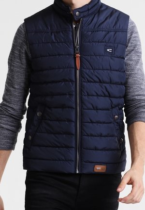 Homme portant un gilet matelassé bleu marine avec une fermeture éclair frontale et des poches à boutons, par-dessus une chemise grise à manches longues, debout devant un fond uni.