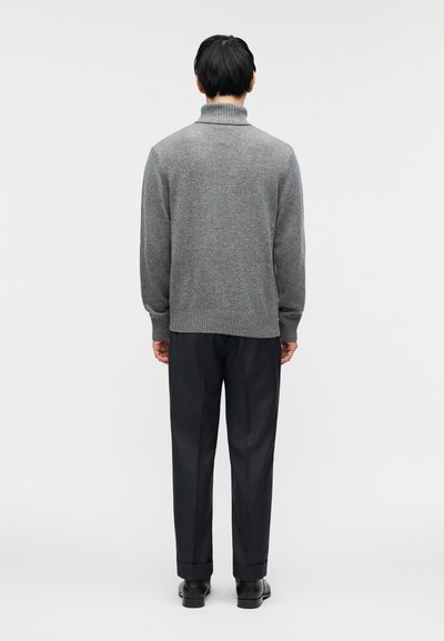 Γκρι πλεκτό πουλόβερ με ψηλό γιακά, συνοδευόμενο από σκούρο παντελόνι με ρίγες, που φαίνεται από πίσω. Λεία υφή με ριμπ λεπτομέρειες.