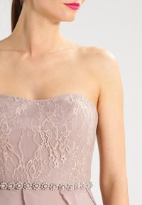 Šaty bez ramínek v jemné mauve krajce s texturovaným vzorem, s páskem doplněným perlami pro zvýraznění detailu.