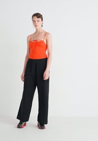 Body aderente arancione senza maniche con cordino frontale, abbinato a pantaloni neri larghi e scarpe da ginnastica rosse e nere. Sfondo bianco semplice.