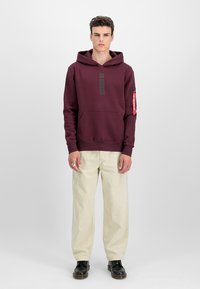 Ung mand iført en vinrød hættetrøje med frontlomme og beige viddebukser, stående oprejst mod en ensfarvet lys baggrund.