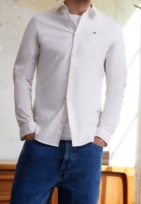 White long-sleeve shirt with a collar, featuring a small logo on the chest, paired with blue denim jeans. Smooth fabric texture.
