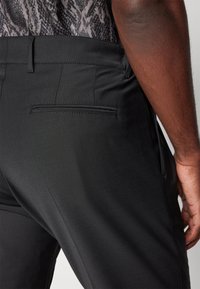 Pantalon noir au tissu lisse, avec une poche arrière et une coupe ajustée. La taille est élégante et structurée pour un look soigné.