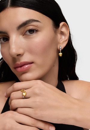 Kobieta o ciemnych włosach, nosząca złoty pierścionek w kształcie serca oraz pasujące złote kolczyki w kształcie serca na srebrnej obręczy, ubrana w czarną bluzkę.