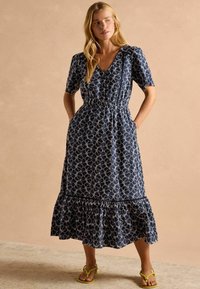 Marineblaues Kleid mit weißem Blumenmuster, kurzen Ärmeln, tailliertem Design und gestuftem Rüschensaum. Kombiniert mit leuchtend gelben Sandalen.