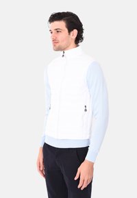 Gilet imbottito bianco con colletto alto, chiusura zip frontale e dettagli neri. Indossato sopra una camicia a maniche lunghe azzurra, abbinato a pantaloni scuri.