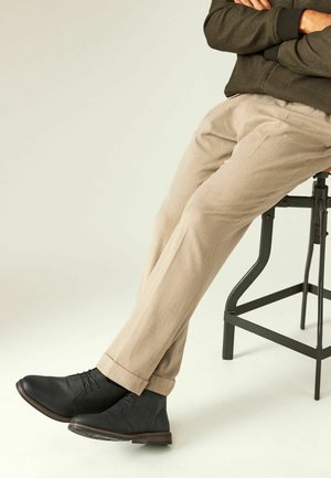 Botines de cuero negro con cordones y ribete marrón, combinados con unos pantalones beiges remangados, sentados en un taburete de metal negro.