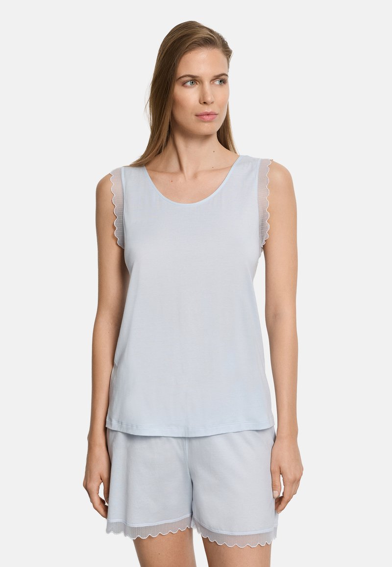 Nainen, joka pukeutunut vaaleansiniseen hihattomaan toppiin ja siihen sopiviin shortseihin, joissa on aaltoilevat pitsireunukset, seisoo valkoista taustaa vasten.