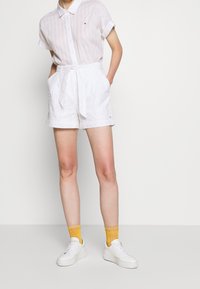 Hvid kortærmet skjorte med knapper, kombineret med hvide shorts med et bundet bælte og blomsterbroderi. Båret med hvide sneakers og gule prikkede sokker.