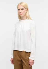 Donna bionda che indossa una camicia bianca a maniche lunghe con colletto e polsini volant, e pantaloni marroni su misura, contro uno sfondo bianco.