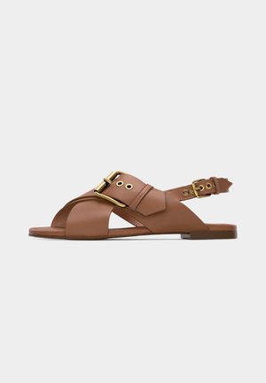 Barna bőrből készült lapos szandál keresztben átlapolt pántokkal és arany csatt díszítéssel, nyitott orral és állítható hátsó pánttal.
