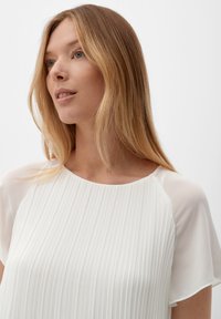 Blouse blanche plissée avec des manches courtes transparentes, un col rond et une texture lisse. Le tissu présente des rayures verticales pour un détail supplémentaire.