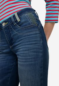 Granatowe dżinsy mają prosty krój, subtelne detale wyblaknięcia, zielone przeszycia oraz dekoracyjny wzór na tylnej kieszeni.