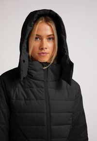 Giacca nera imbottita con colletto alto e cappuccio regolabile. Presenta una texture liscia e una trapuntatura orizzontale. Chiusura con zip e bottoni a pressione.