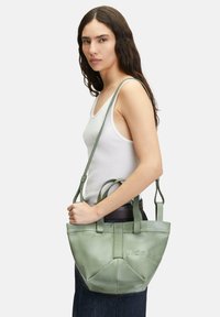 Zielona skórzana torba na ramię z podwójnymi uchwytami i odpinanym paskiem na ramię, z geometrycznym wzorem i wytłoczonym napisem.