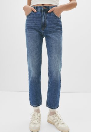 Person iført højtaljede blå jeans med hænderne i lommerne, kombineret med hvide sneakers og lyse sokker mod en ensfarvet baggrund.