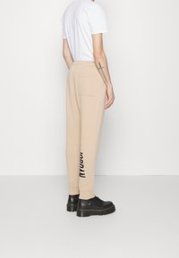 Beige sweatpants med avsmalnande ben, med vit logotryck och en bakficka, bärs med en vit T-shirt och svarta platåskor.